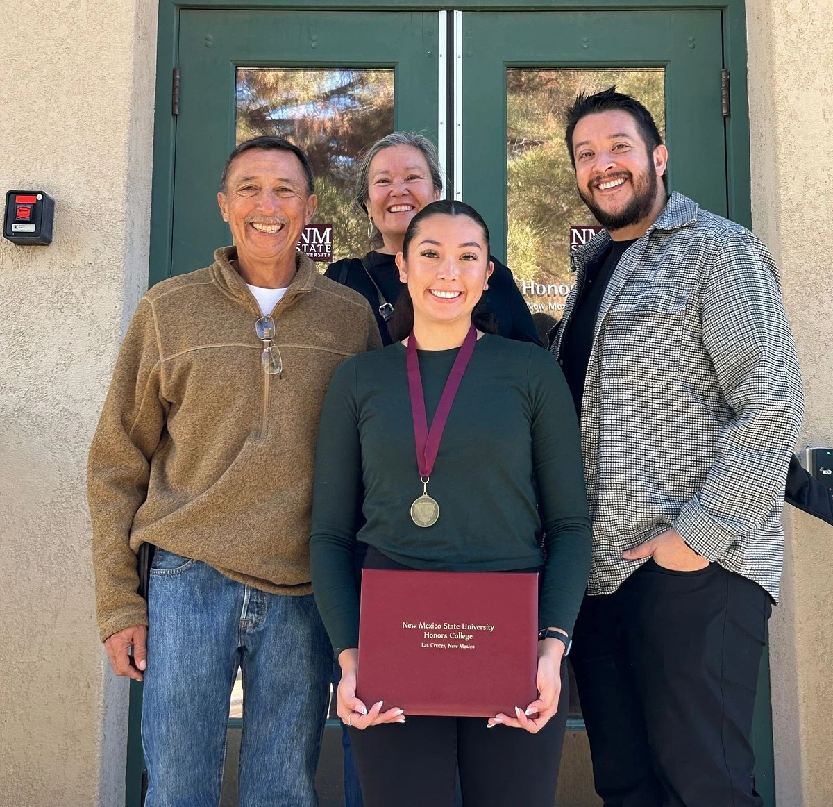 A young woman holding a New Mexico State University Honors College certificate poses with three smiling adults in front of the Honors building.